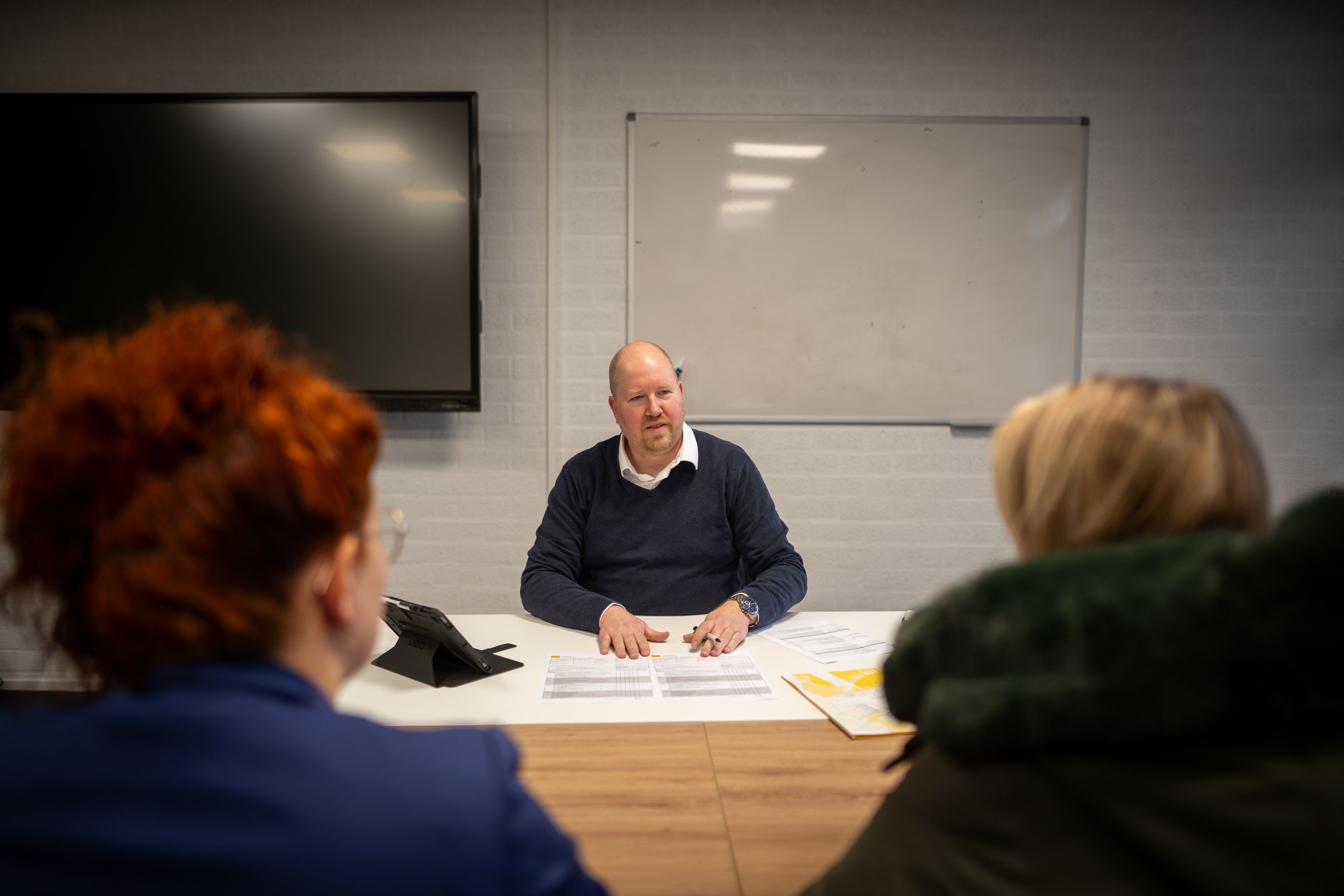 Een man zit met zijn handen op de tafel achter wat documenten. Twee vrouwen zitten voor de tafel.