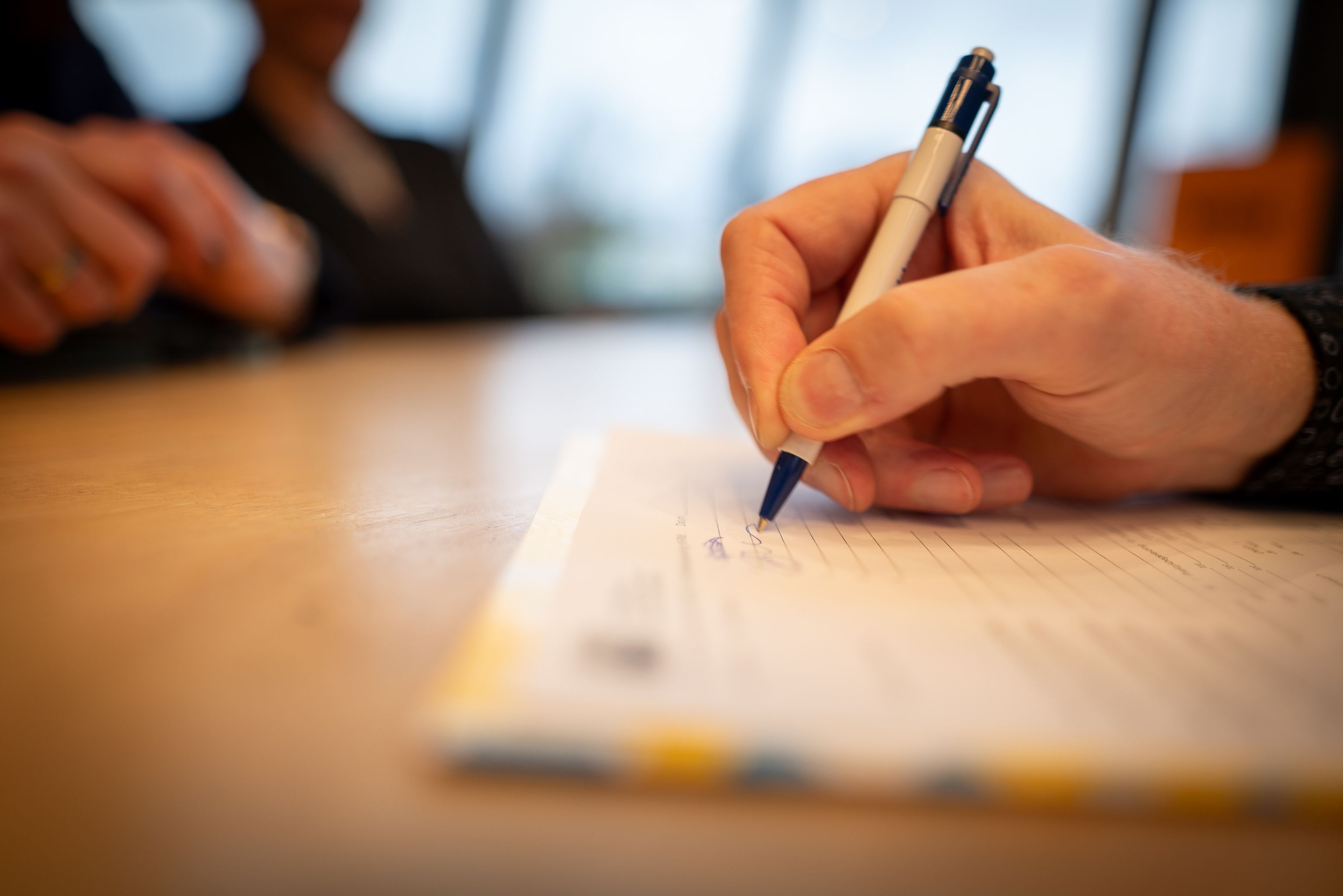 Een foto van een hand van een oudere man die een formulier invult op een tafel. Aan de overkant zie je de handen van de mensen aan de andere kant van de tafel.