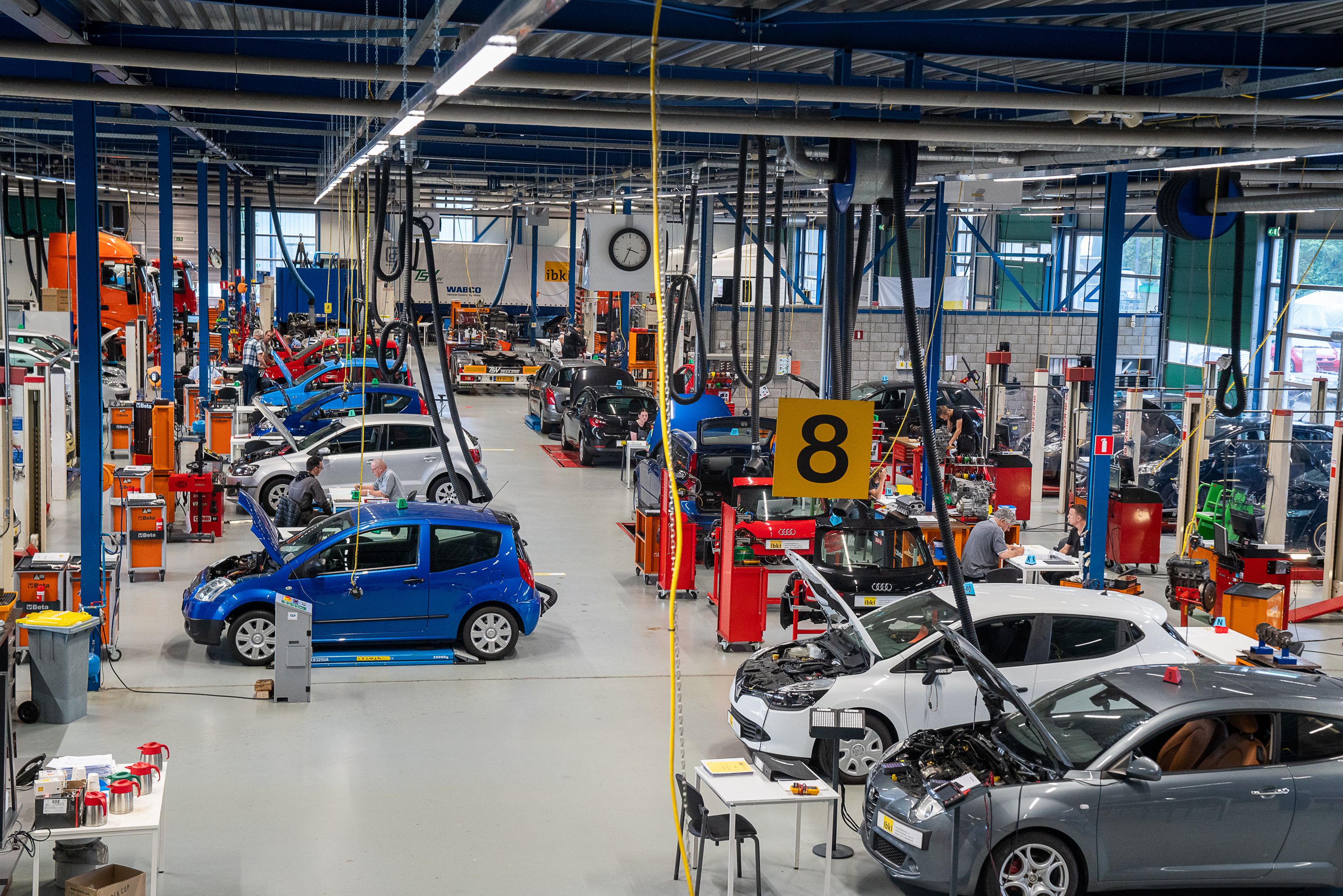Overzicht van de examenhal met opstelling van auto's en een geel bord met het cijfer 8.