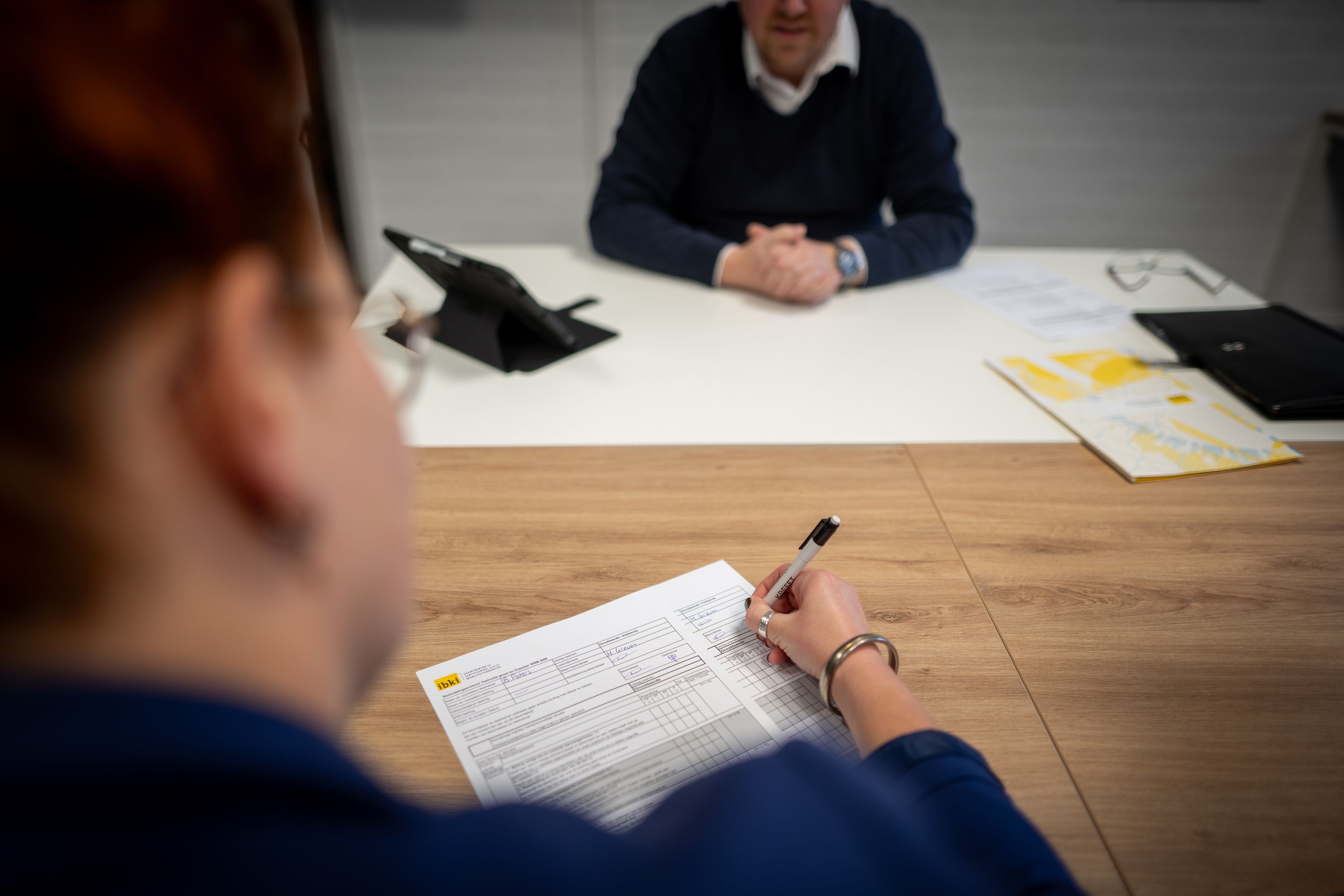 Een vrouw vult een formulier in op een tafel. Op de achtergrond zit een man met zijn handen ineen achter de tafel.
