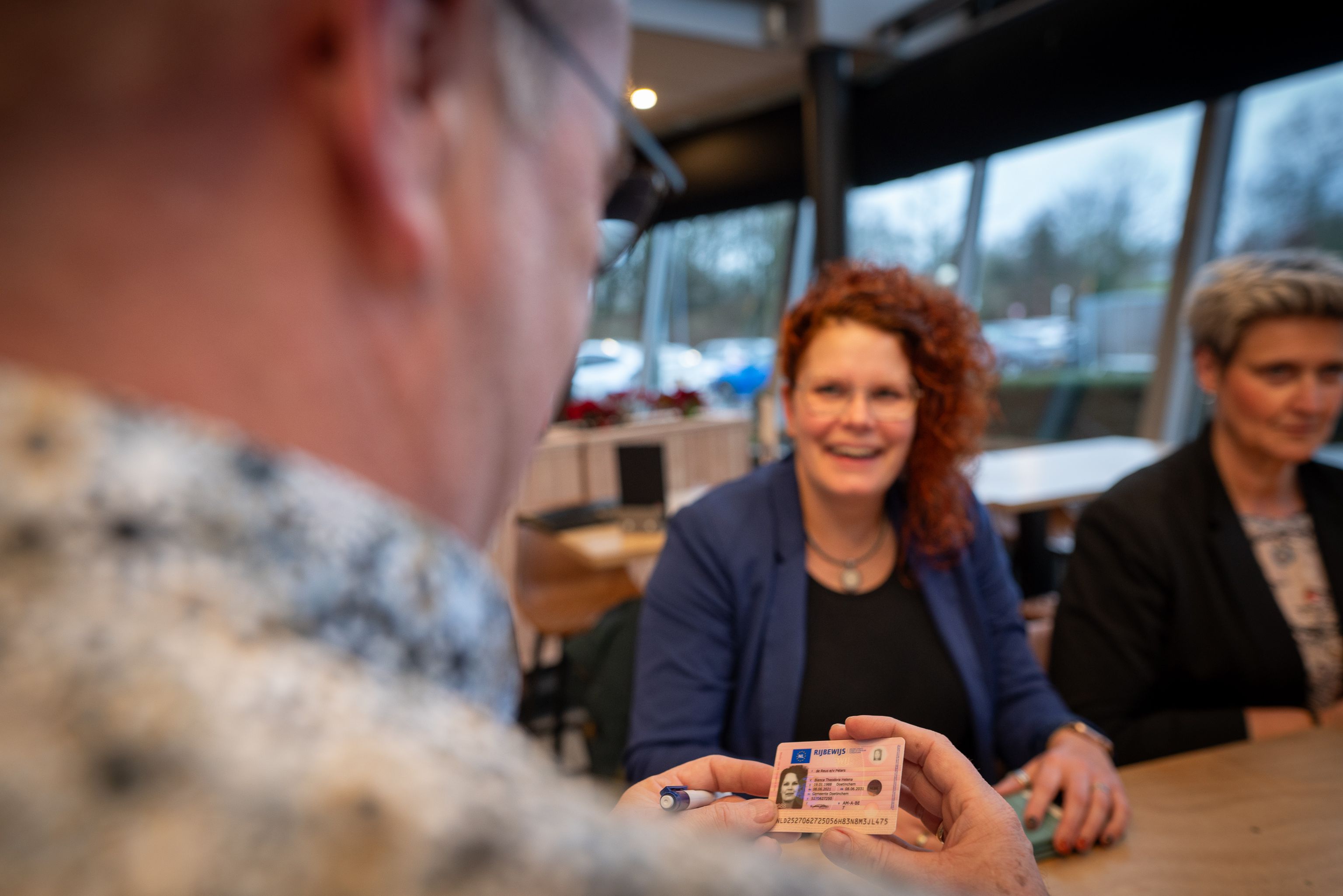 Een examinator controleert het rijbewijs van een deelnemer. Hij zit aan een tafel met twee vrouwen.