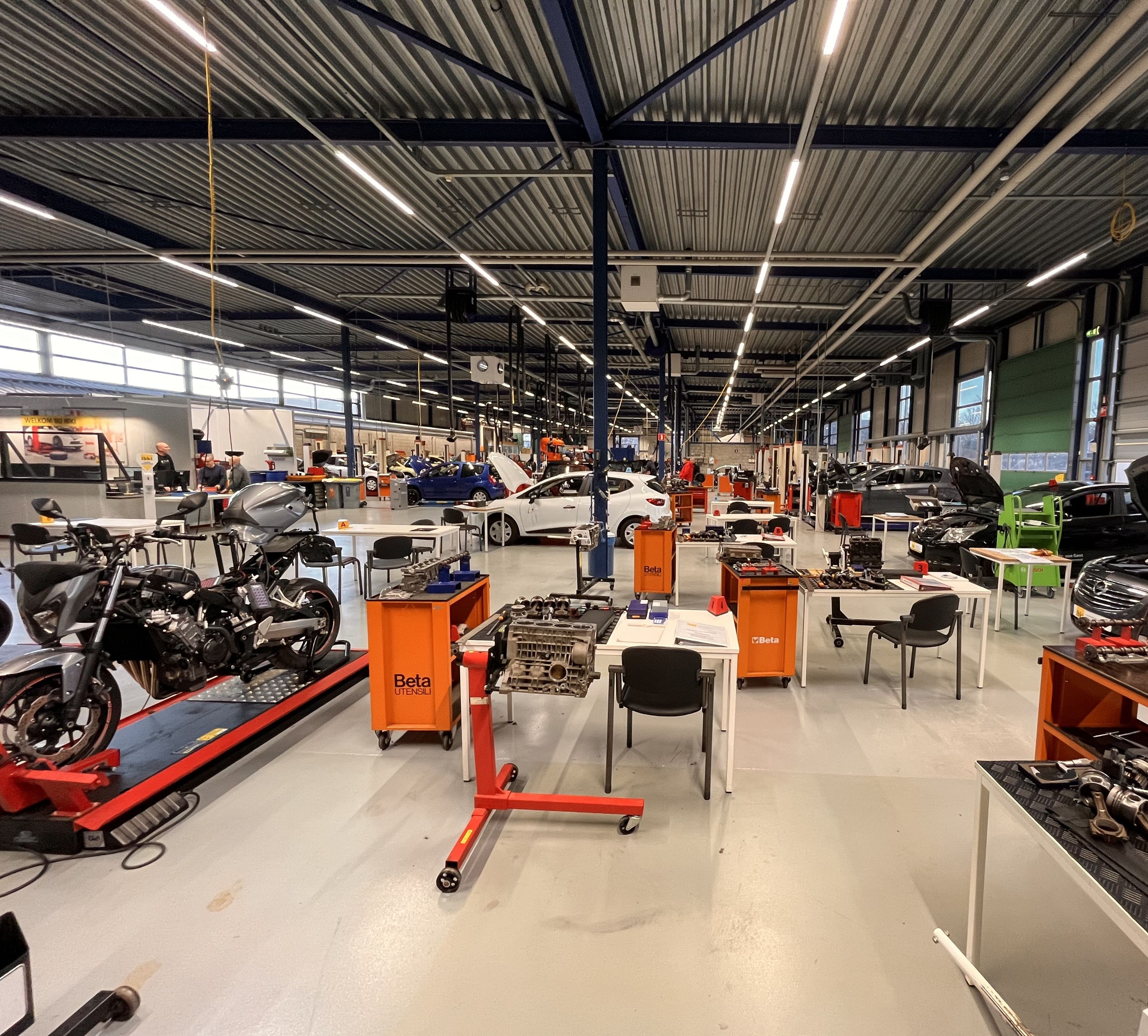 Zicht op de examenhal van IBKI. Links staat een motorfiets. Er staan gereedschapskarren en op de achtergrond auto's. Er staan ook tafels en stoelen.