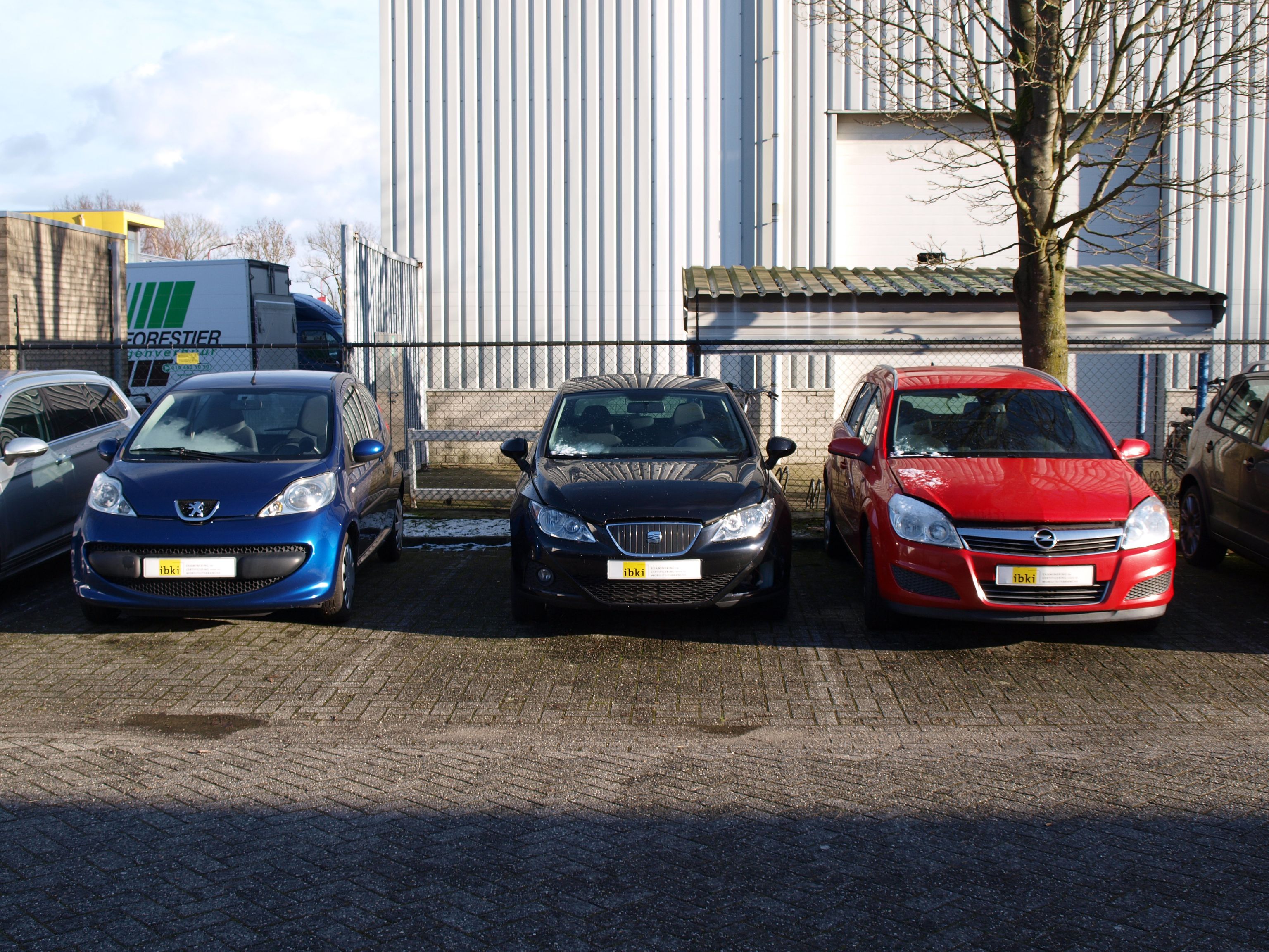 Drie auto's, met een nummerplaat van IBKI, naast elkaar op een parkeerplaats. Op de achtergrond een gebouw en een stukje van een vrachtauto.