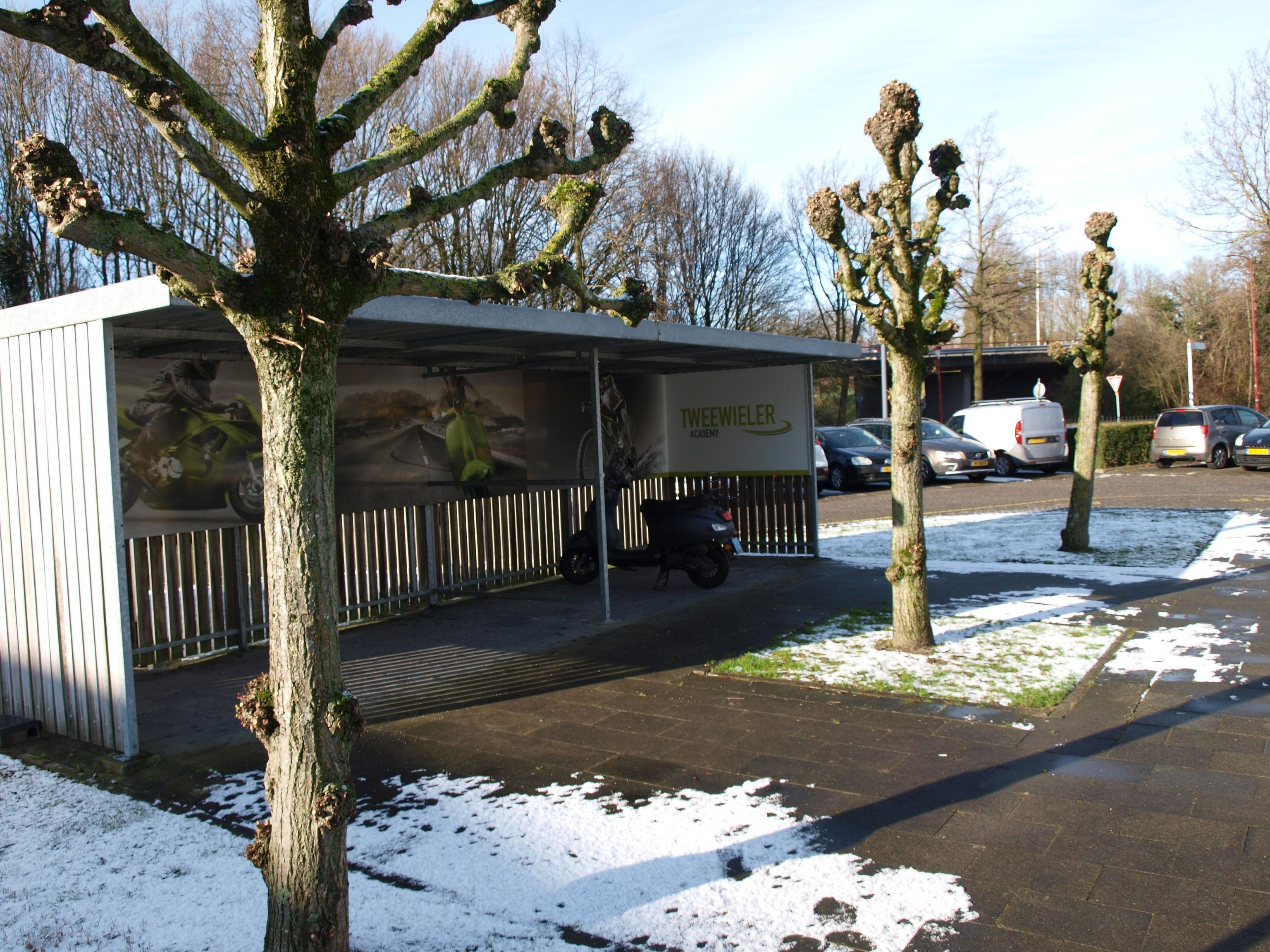 Er staat een scooter onder een overkapping. Daar achter is de parkeerplaats met auto's.