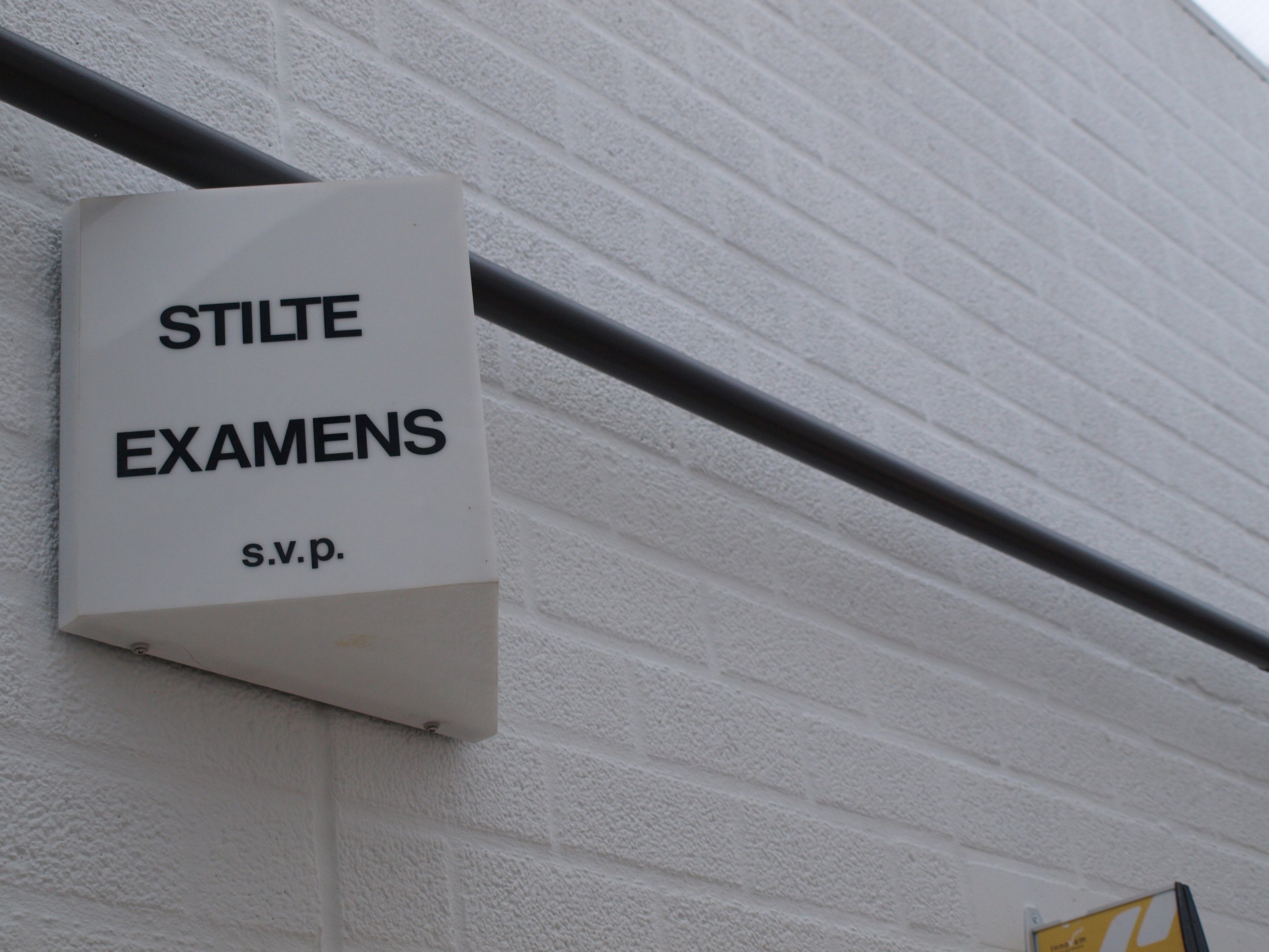 Een lamp aan een muur met daarop 'Stilte examens s.v.p.'. De muur is wit met een zwarte streep.