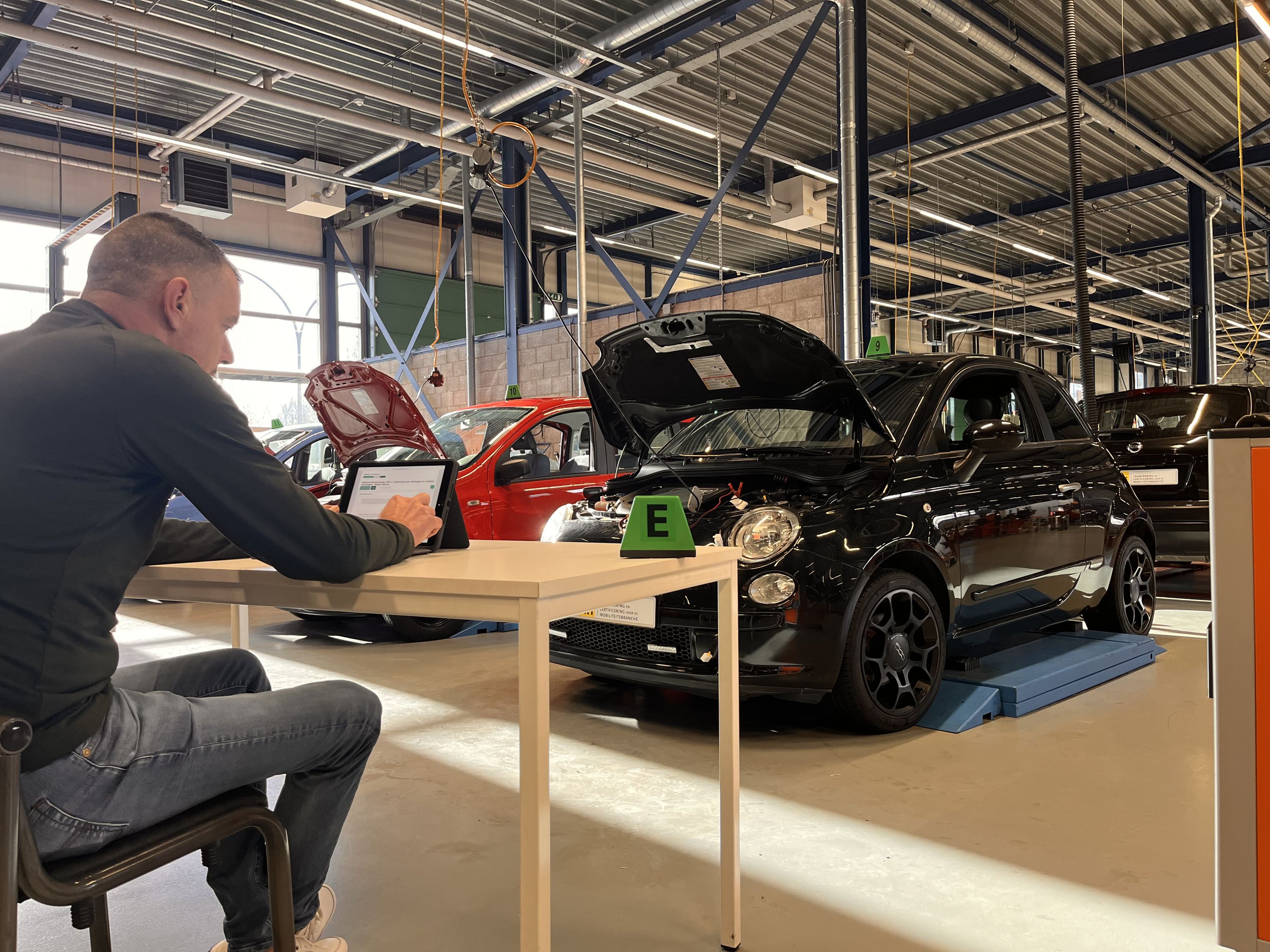 Een deelnemer aan het examen achter een iPad. Twee auto's met open motorkap.