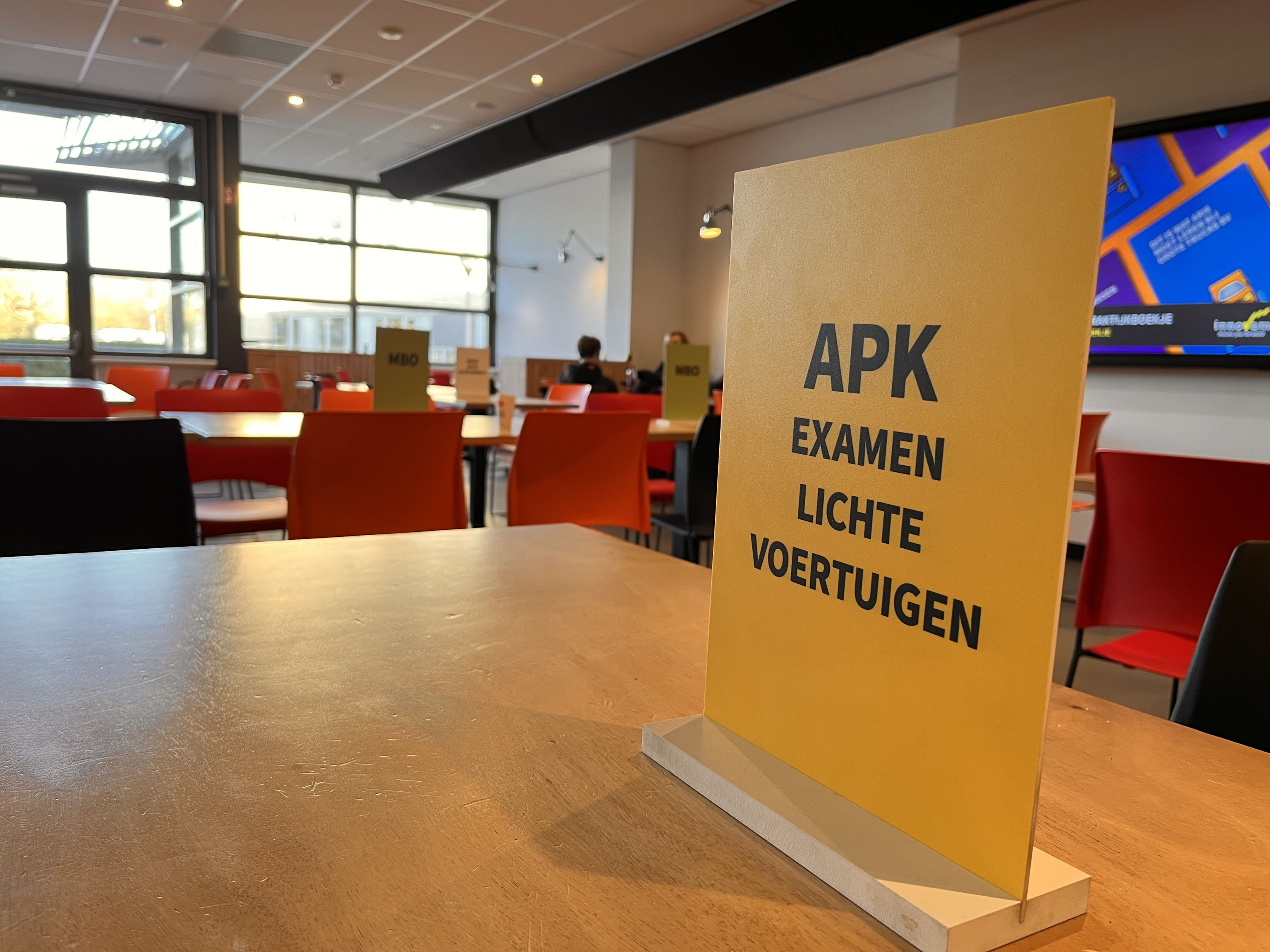 Op tafel staat een geel bord met daarop 'APK examen lichte voertuigen'. Op de achtergrond meer tafels en stoelen.
