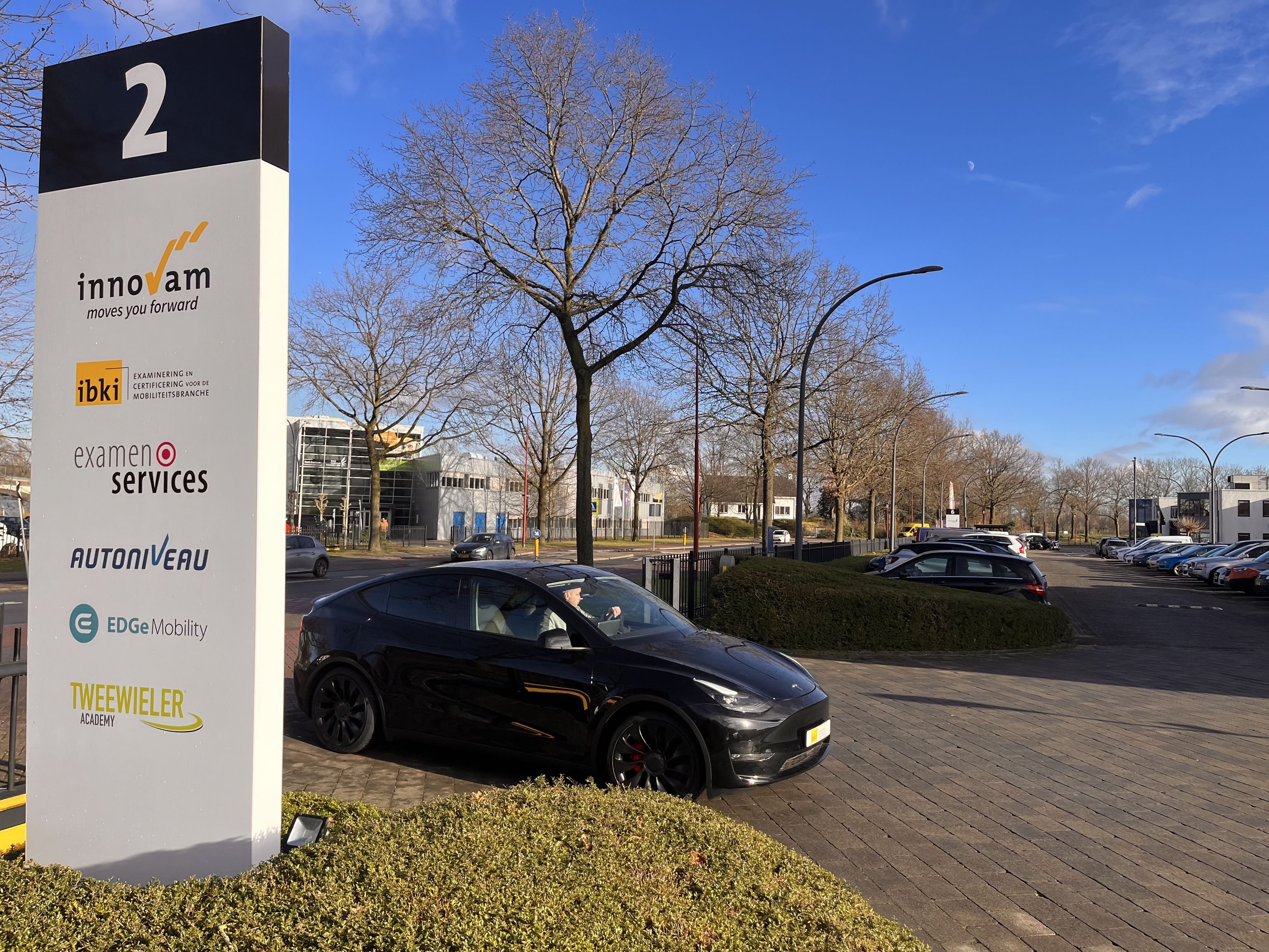 Het parkeerterrein van IBKI. Op de hoek is het naambord van nummer 2 te zien. Daarachter de weg waar auto's rijden.