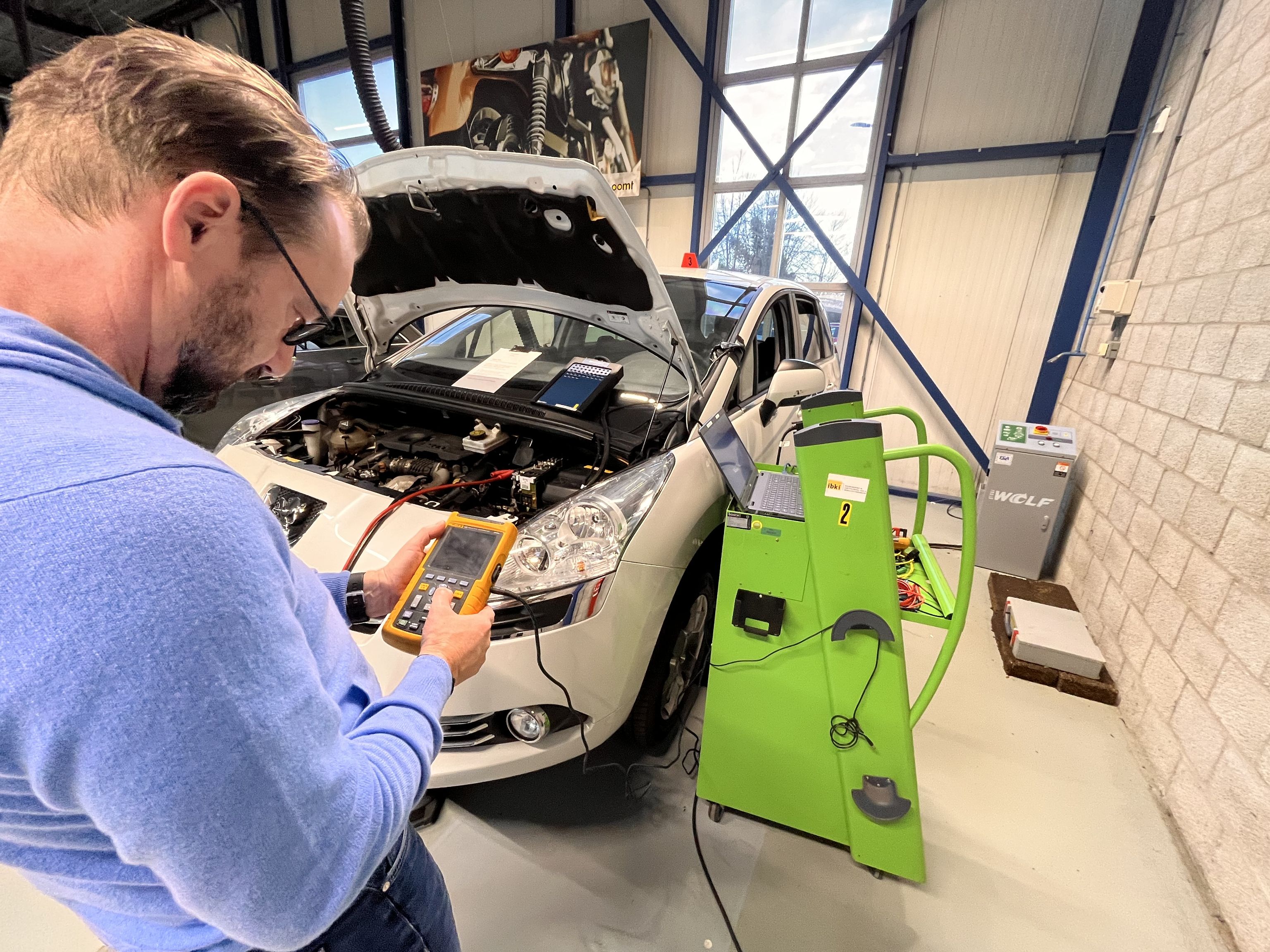 Een man staat met een multimeter in zijn hand bij een personenauto. De motorkap staat open en er wordt iets gemeten.