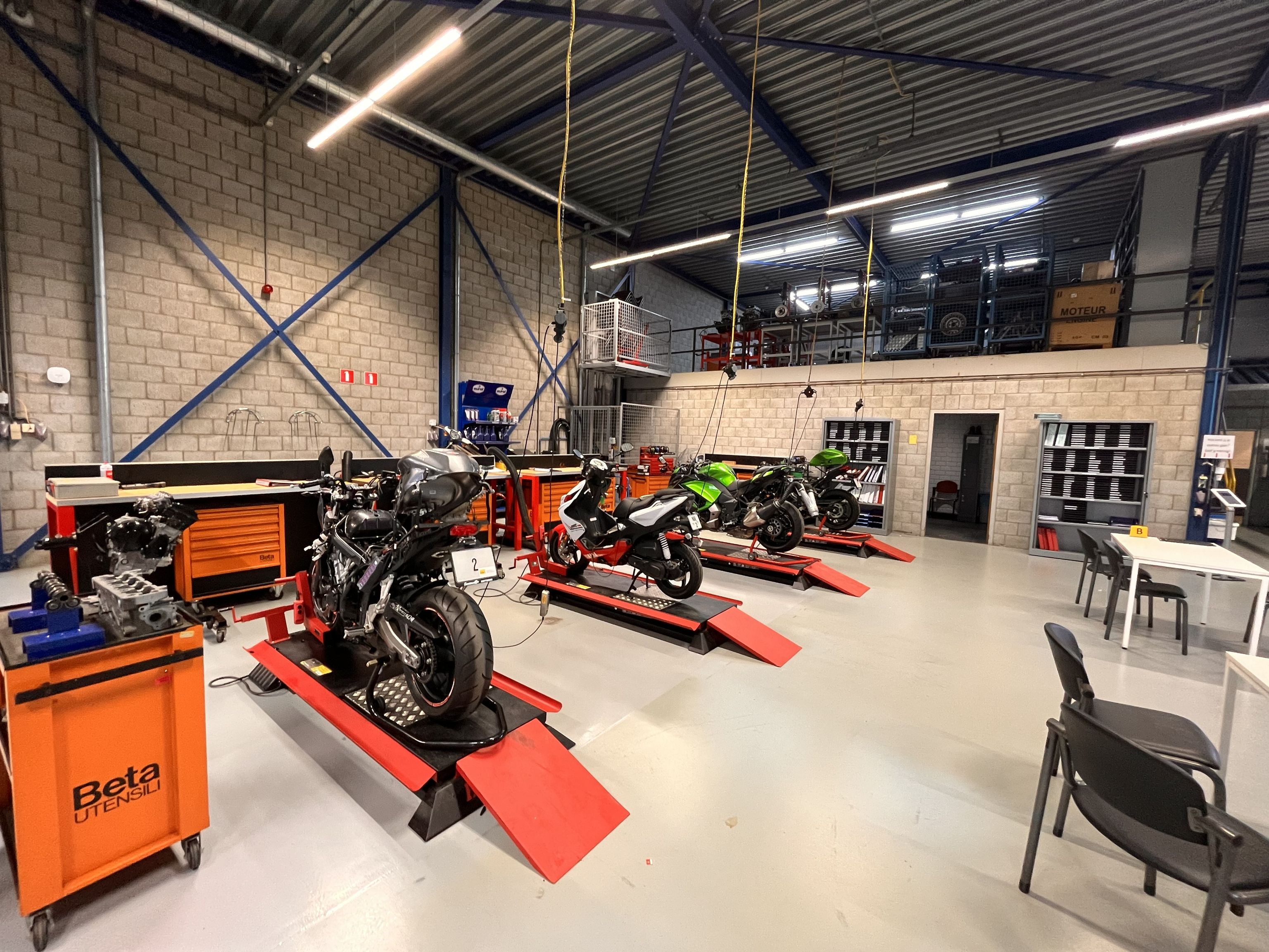 Motorfietsen die op motorheftafels staan in de examenhal. Er staan ook tafels, stoelen en gereedschapskarren.