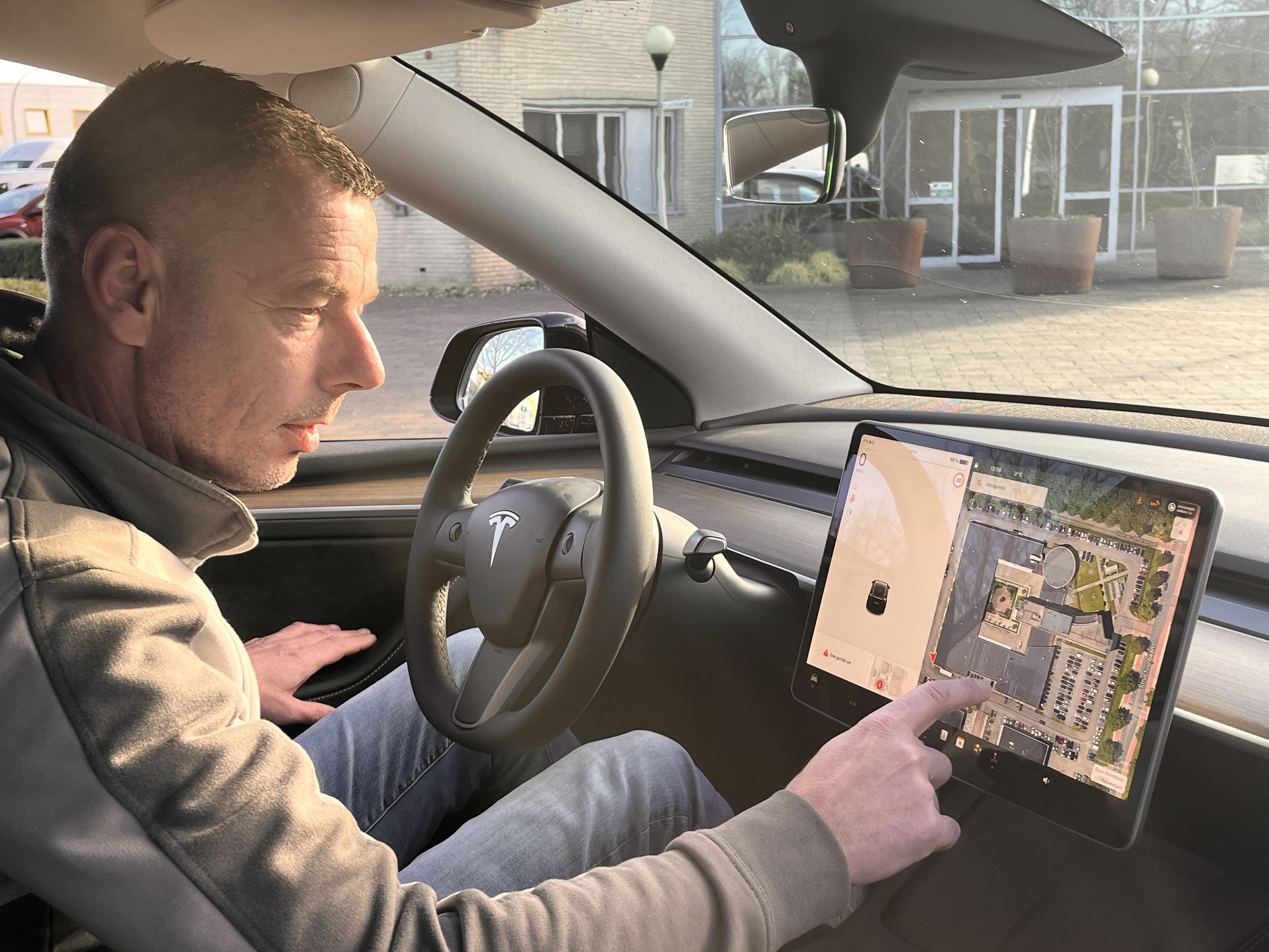Een man in een personenauto tikt met zijn wijsvinger op het navigatiesysteem. Op de achtergrond is de ingang van een gebouw.