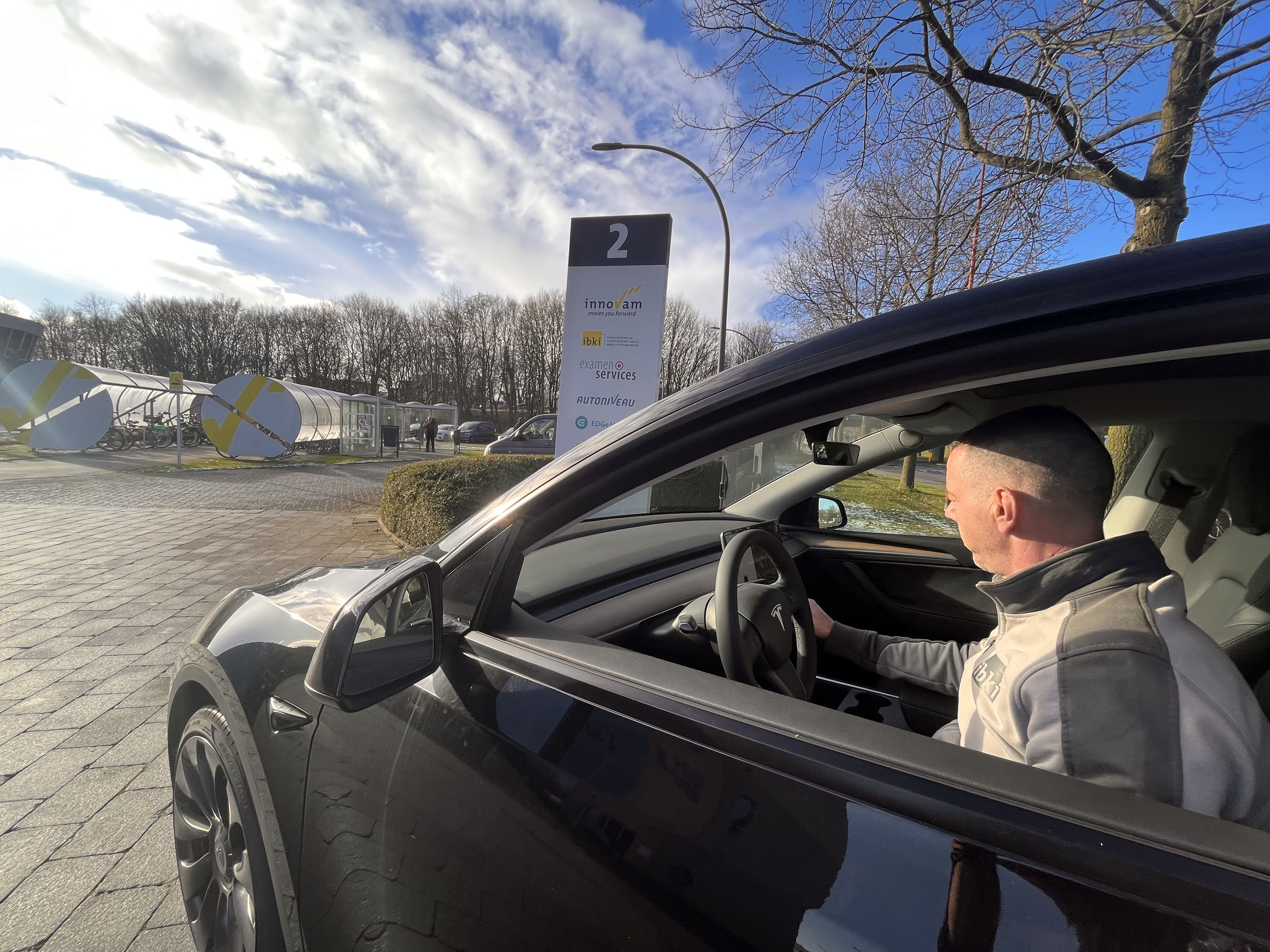 Een man achter het stuur van een personenauto. Op de achtergrond het naambord en de fietsenstalling van het bedrijf.