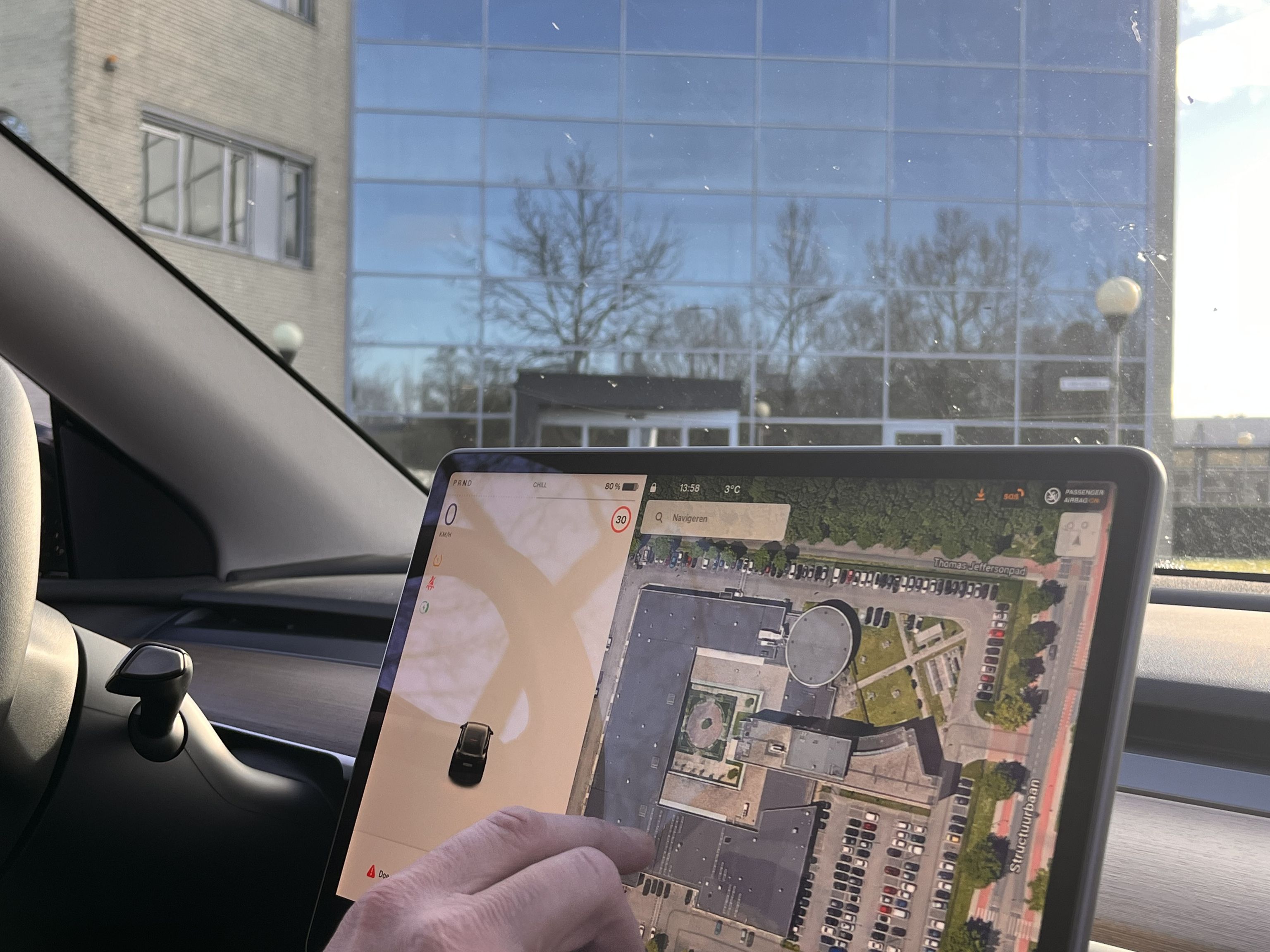 Een hand van iemand die met het navigatiesysteem in zijn auto bezig is. Op de achtergrond de weerspiegeling van bomen in de ramen van een gebouw.