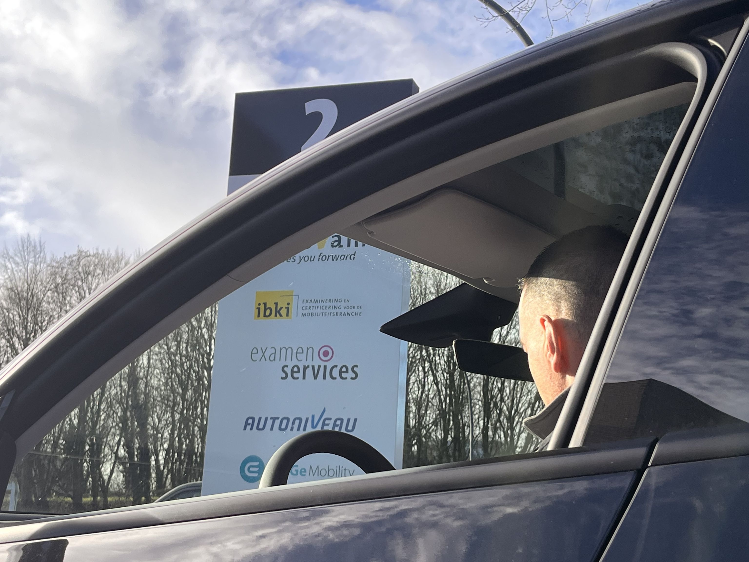 Een man zit achter het stuur van een auto. Door het raam zie je het naambord waar o.a. IBKI op vermeld staat.