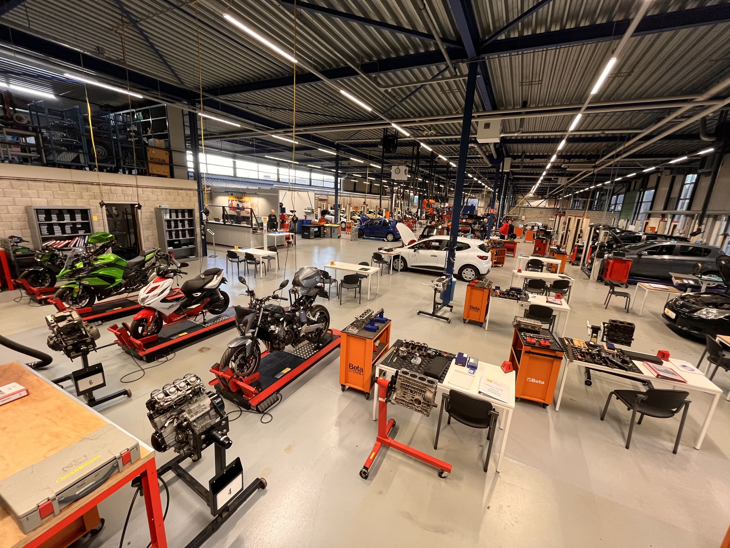 De examenhal van IBKI met op de voorgrond motorfietsen en tafels waaraan je examen kunt doen. Verderop staan personenauto's en gereedschapskarren.