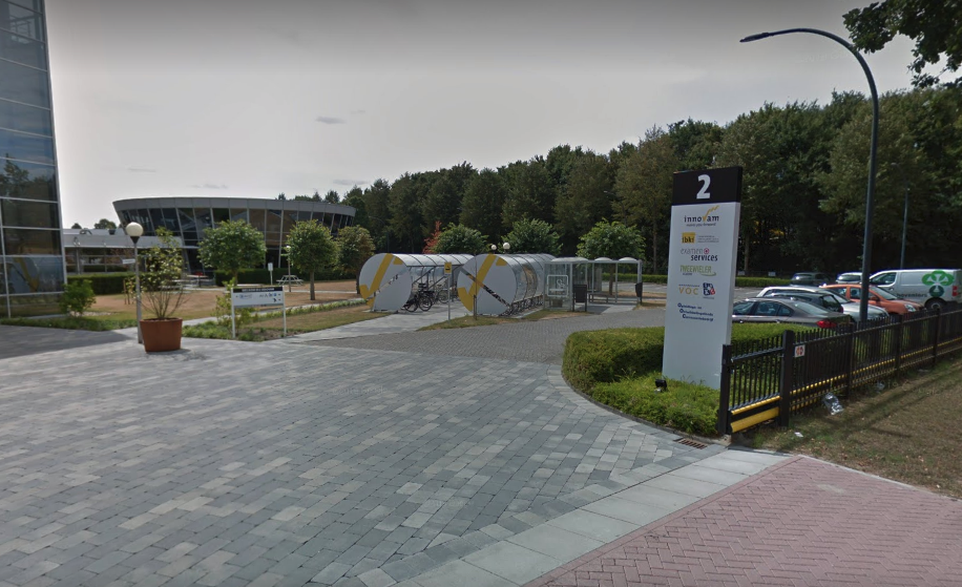 De ingang van het terrein van IBKI met parkeerplaats en fietsenstalling. Op de achtergrond staan bomen.